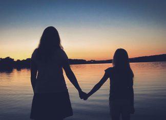 A silouette of a woman and young girl are seen, facing away from camera towards a sunset in the distance. They are holding hands.