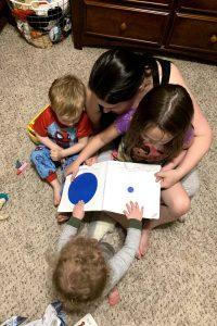 Photo of a mother and three young children sitting on a carpeted bedroom floor. Two children are sitting in the mother's lap and one child is sitting in front of the mother. The mother has a book opened on her lap and all three children are looking at the book.