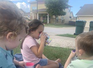 Photo of three young children sitting outside on a blue blanket, drinking out of plastic cups. The book, "Sarah, Plain and Tall" is opened next to them and is being read aloud by the mother, who is not in the photo.