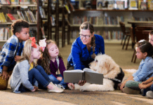 An adult woman wearing a blue sweater in a library reading to a small group of diverse children and a service animal