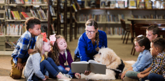 5 Ways to Teach Your Children to be Inclusive An adult woman wearing a blue sweater in a library reading to a small group of diverse children and a service animal