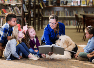 An adult woman wearing a blue sweater in a library reading to a small group of diverse children and a service animal