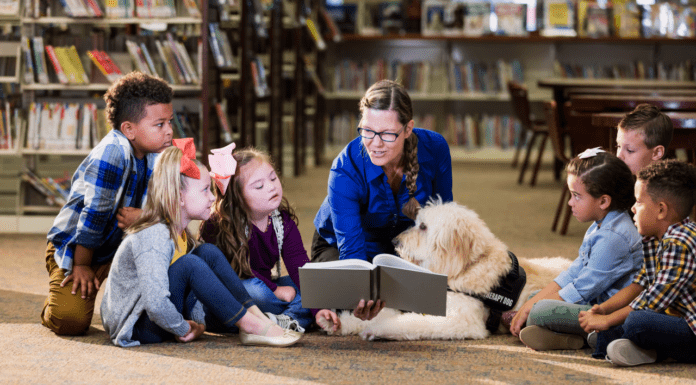 An adult woman wearing a blue sweater in a library reading to a small group of diverse children and a service animal