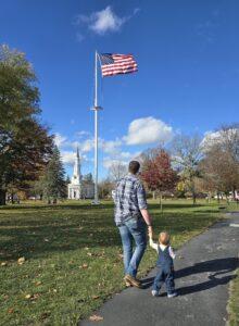 Father and son walking on Lexington Green, church and American Flag in background