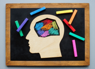 What Autism Is By Identifying What It Isn’t Wooden profile of a head with the area of the brain cut out, laying on a chalkboard. Inside the head, there are multiple colors scribbled to indicate the brain to indicate how different brains function, especially autistic brains.