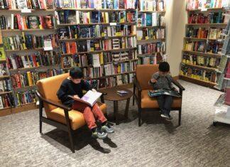 Improving Your Kids’ Literacy Begins at Home Picture of two young boys sitting and reading books in a library, using the easy tips to improve literacy.